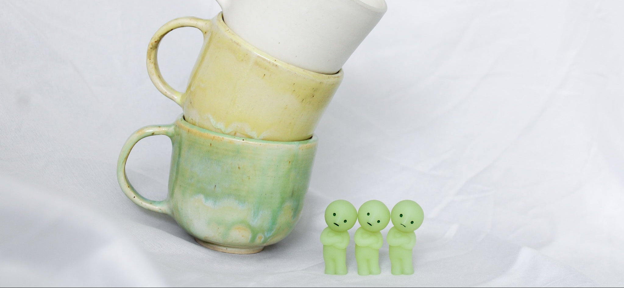 Stack of ceramic mugs in green, yellow, and white on a white background with three small green figures.